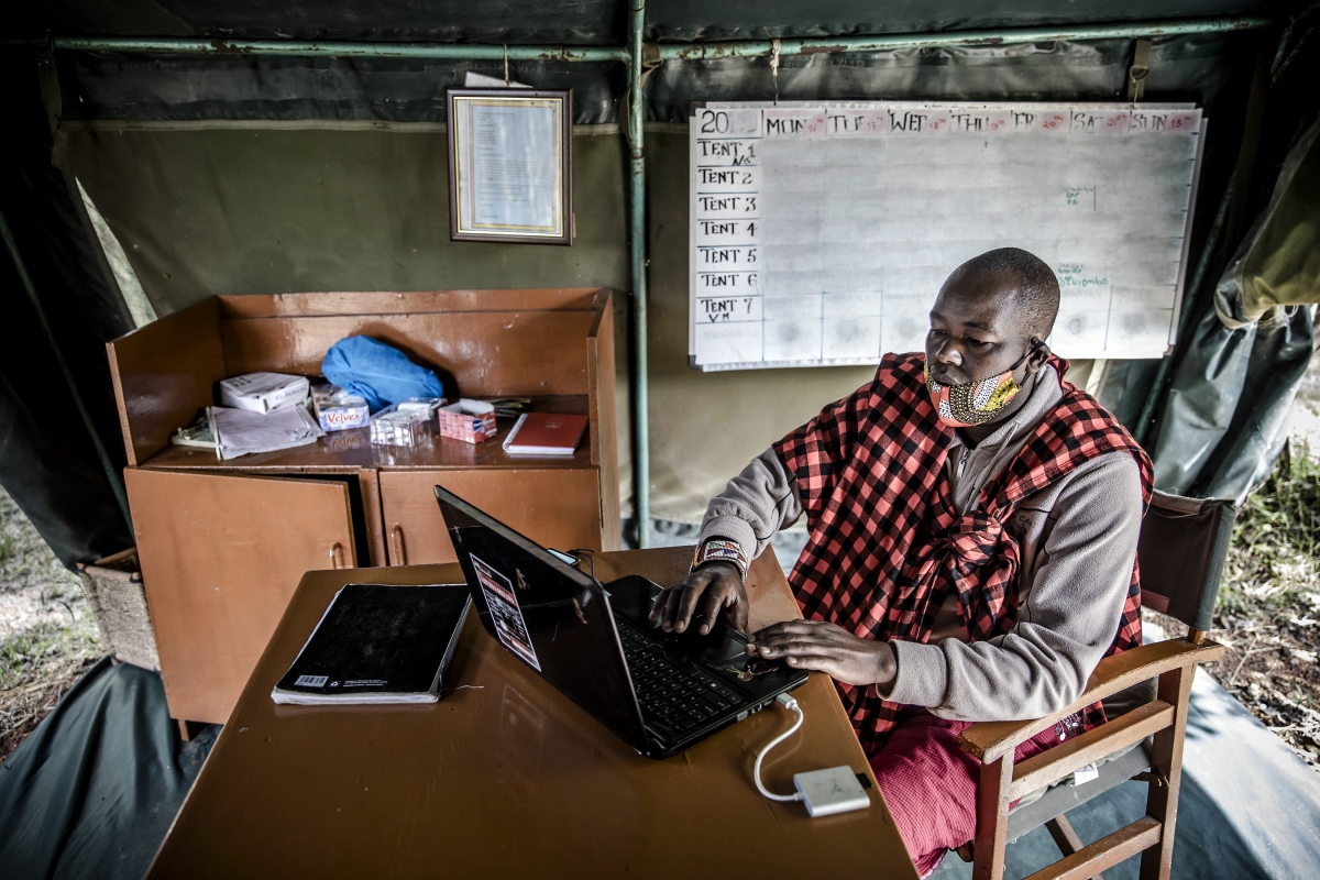 Jimmy Lemara works on his laptop at the Porini Mara Camp, where a blank board that would normally include a list of tourists hangs behind him. MUST CREDIT: Photo for The Washington Post by Luis Tato